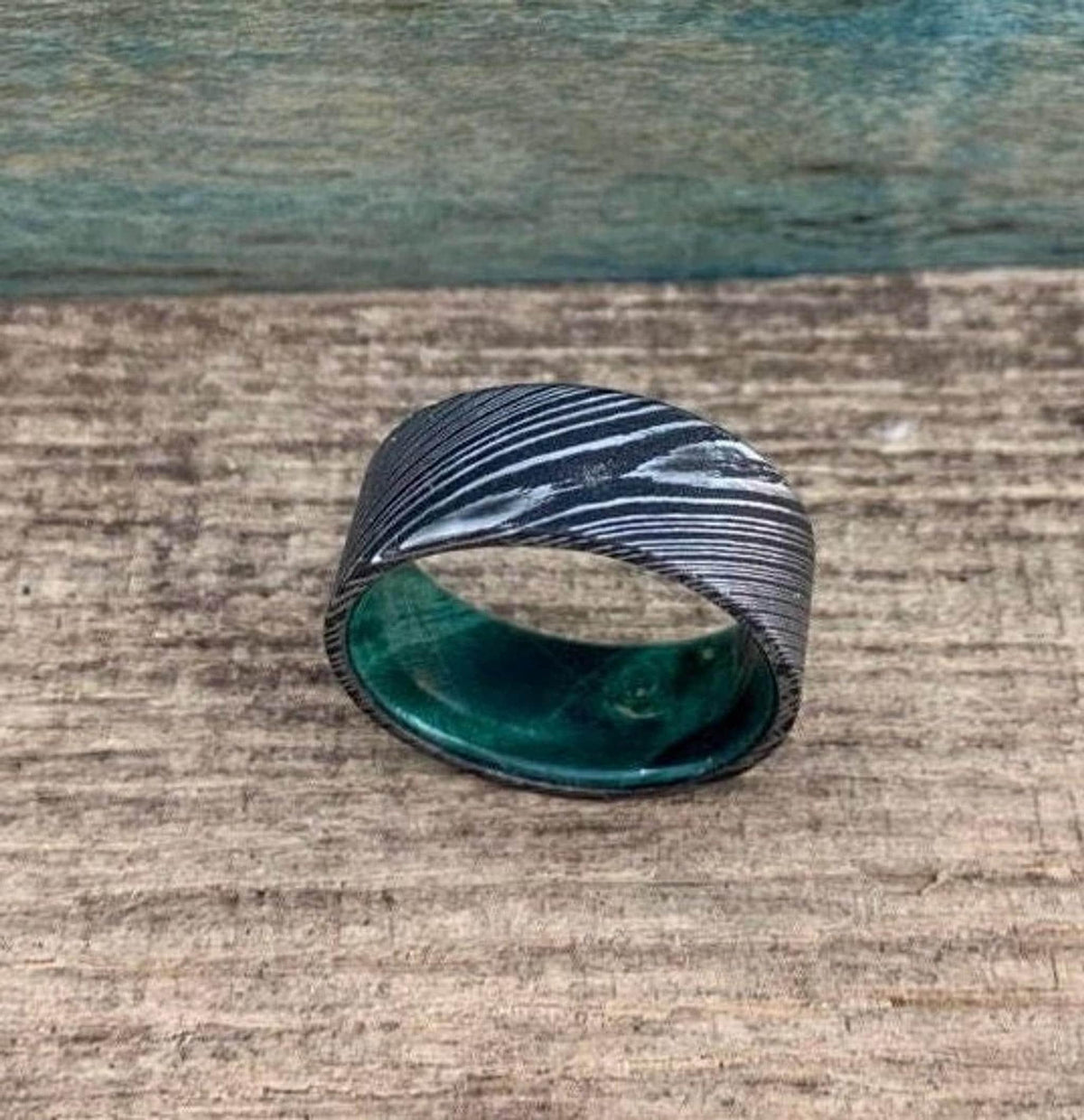 A wide, flat, patterned metal ring with a bright green interior is shown on a wooden surface.