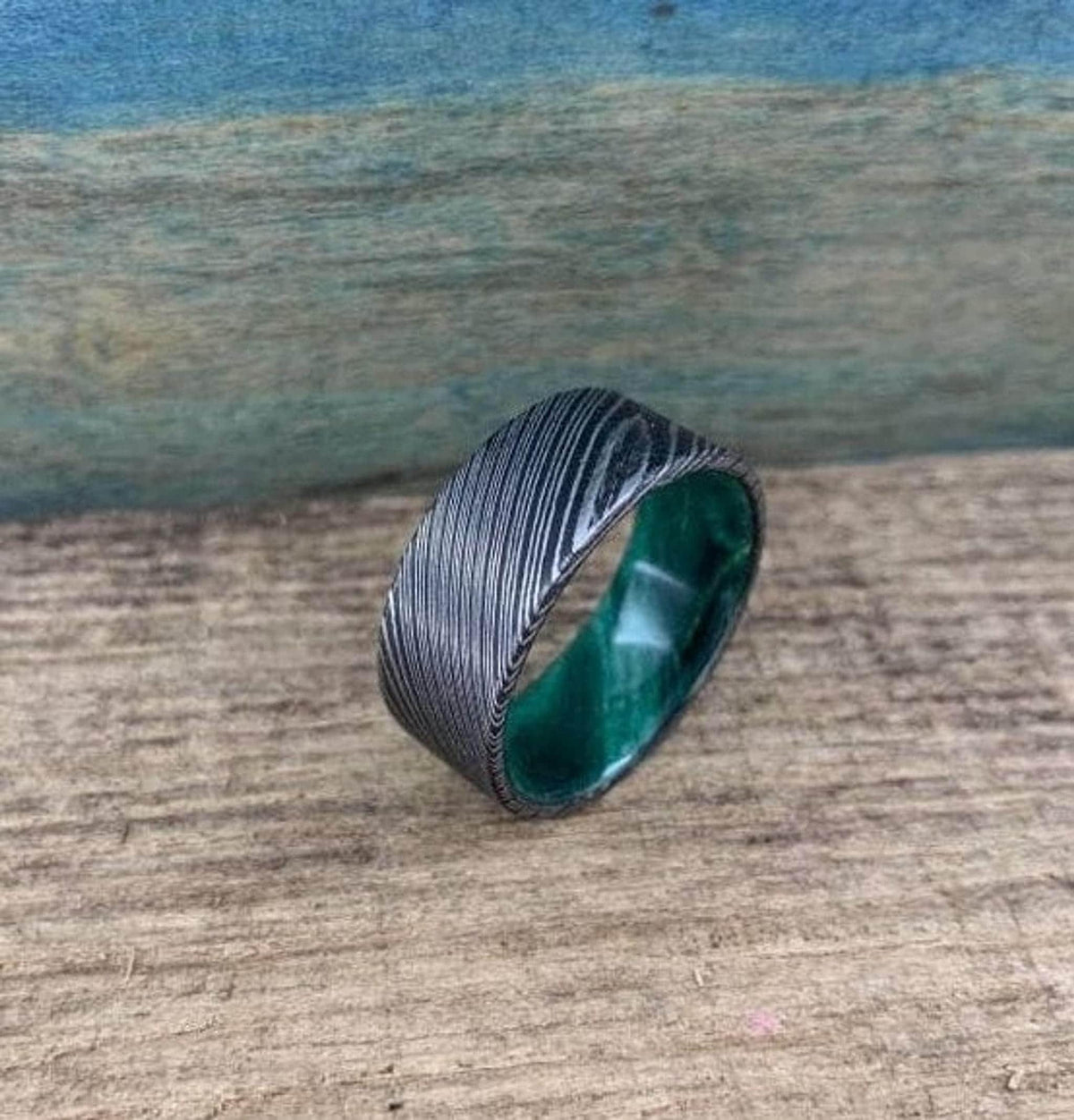 A wide, flat, patterned metal ring with a bright green interior is shown from a slightly different angle on a wooden surface.