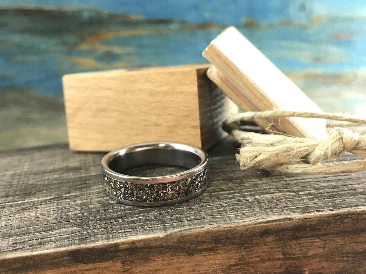 A silver-colored ring with a dark, textured inlay band sits on wood, with two small wooden blocks and rope in the background.