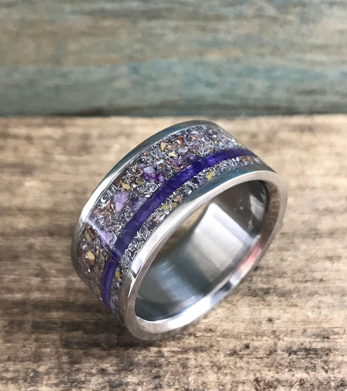 A silver-colored ring with a central inlay of purple and yellow flecks, divided by a thin purple stripe, is shown at an angle on a wooden surface.