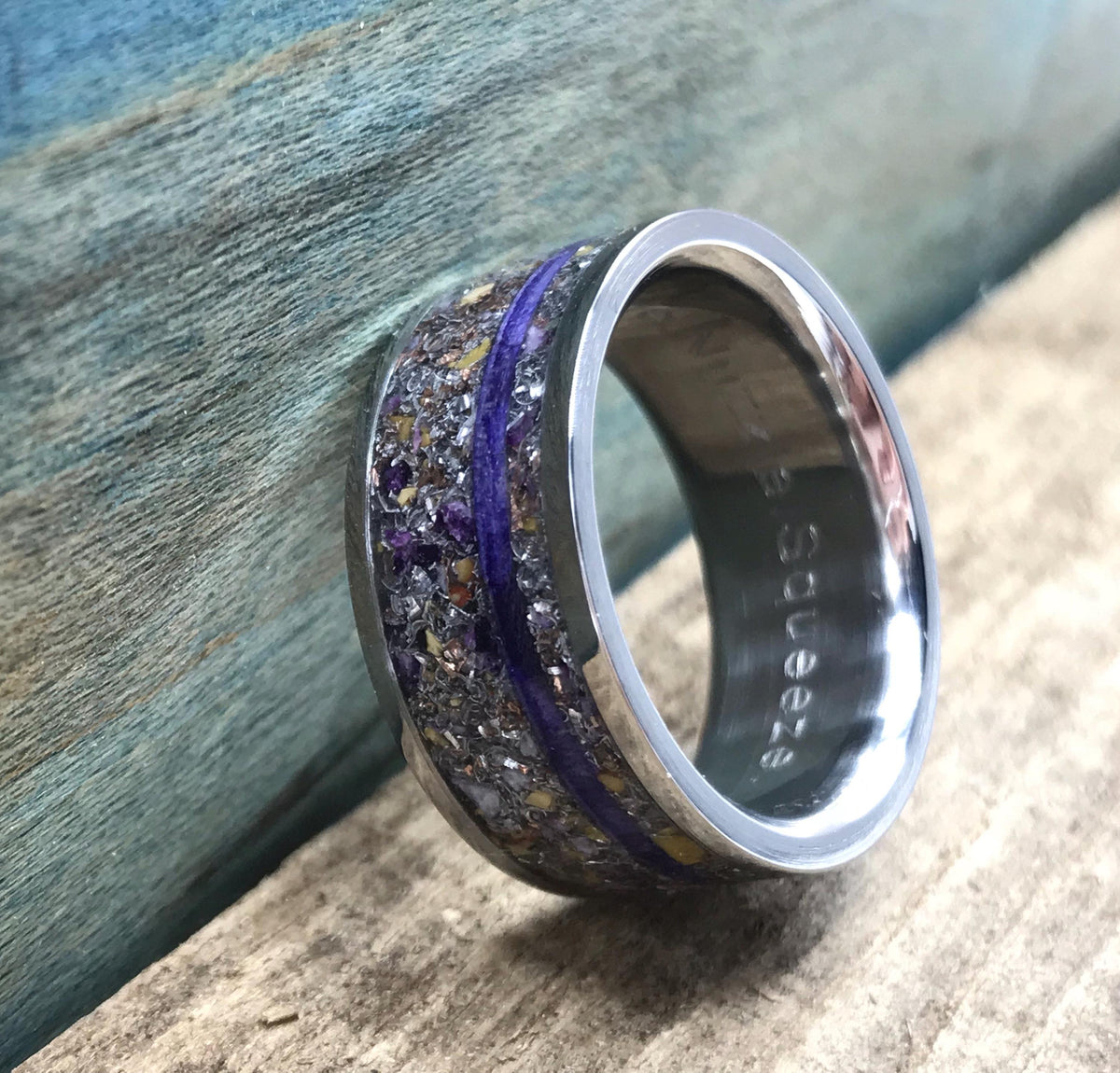 A silver-colored ring with a central inlay of purple and yellow flecks, bisected by a thin purple line, is shown at a steep angle on a wooden surface.