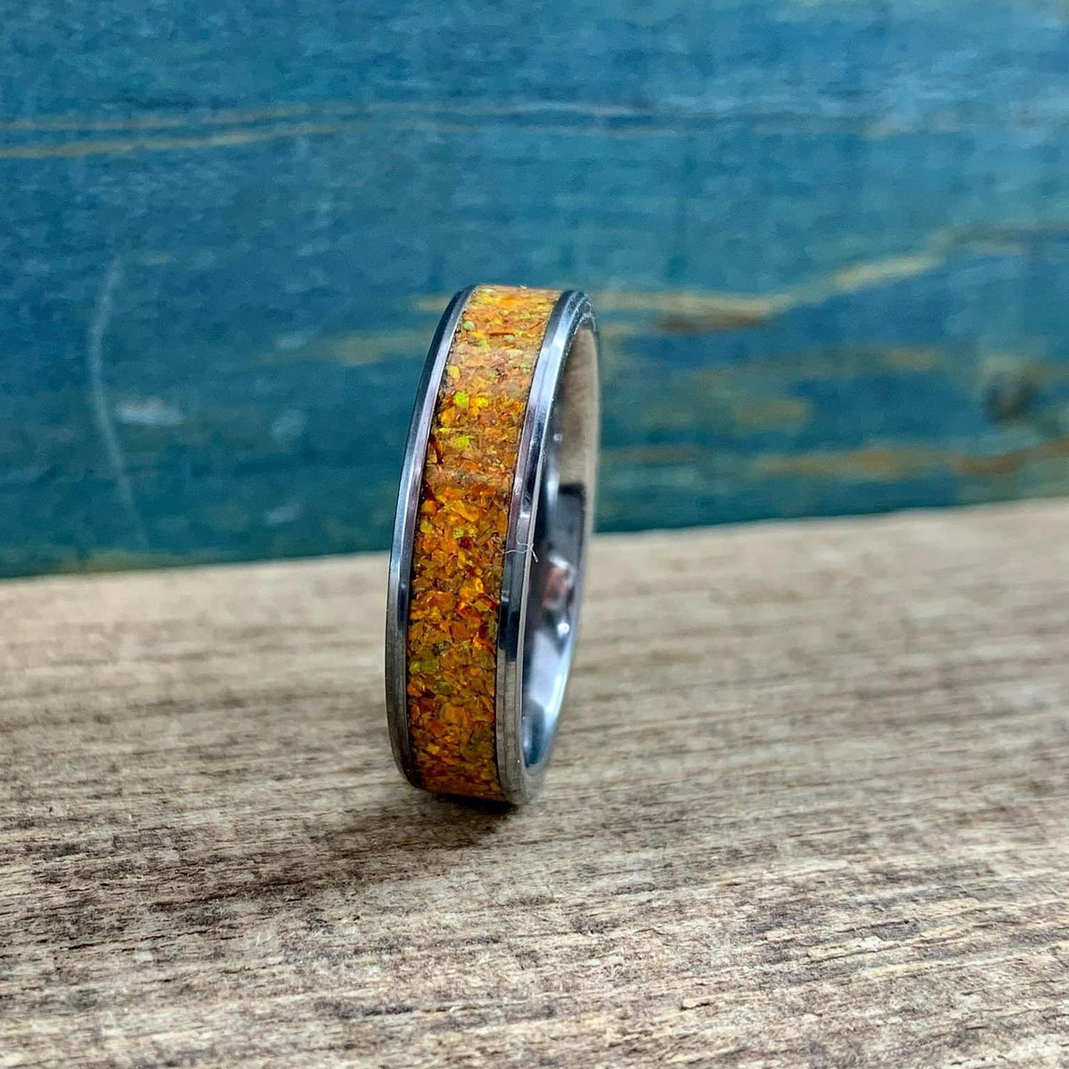A silver-toned ring with an orange glittery inlay, standing upright on a wooden surface with a blue background.