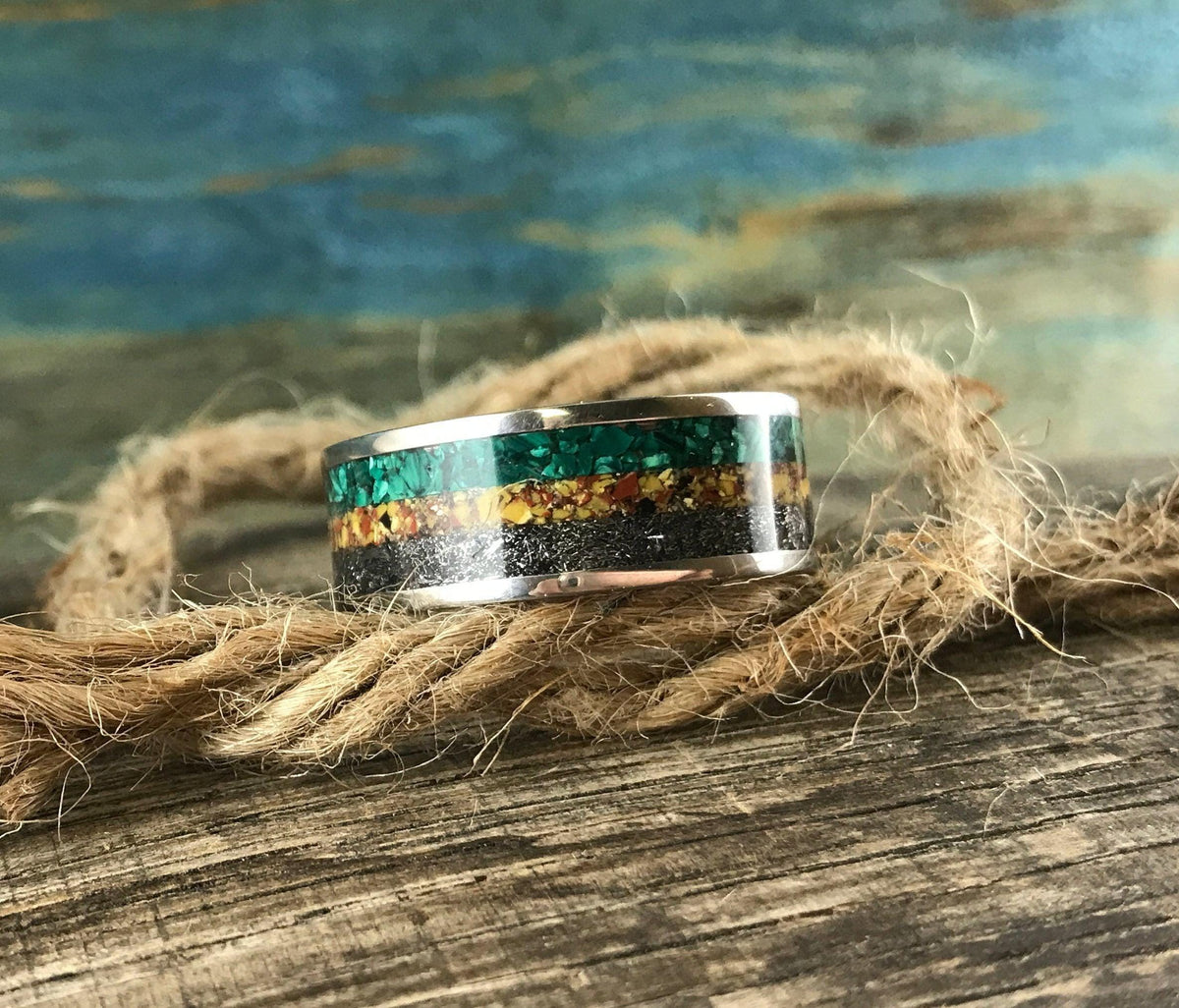A silver ring with green, yellow, and black inlay bands is placed on a coil of thick brown rope.