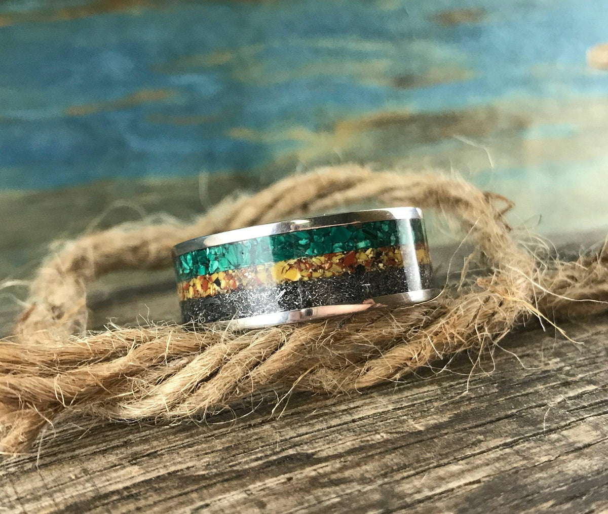 A silver ring with green, yellow, and black inlay bands is centered on a coil of thick brown rope.