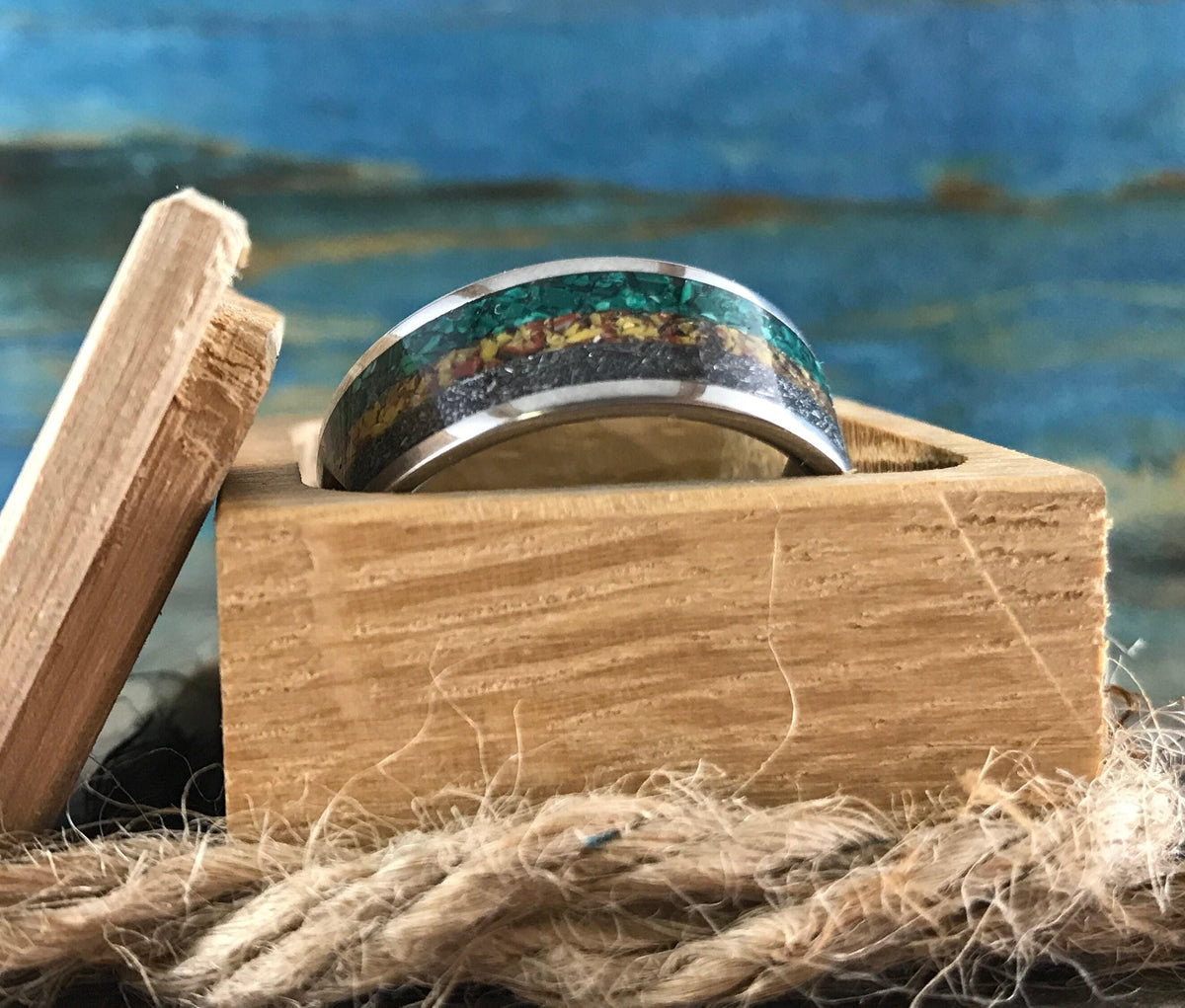 A silver ring with green, yellow, and black inlay bands is nestled inside an open light brown wooden ring box.