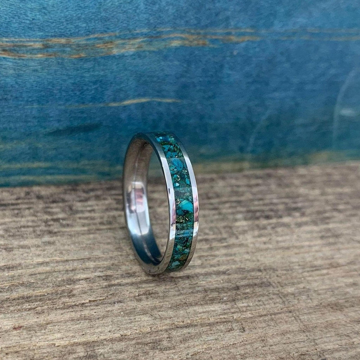 A silver-colored ring with a wide inlay of crushed turquoise and gold-colored flecks, shown at an angle on a wooden surface.