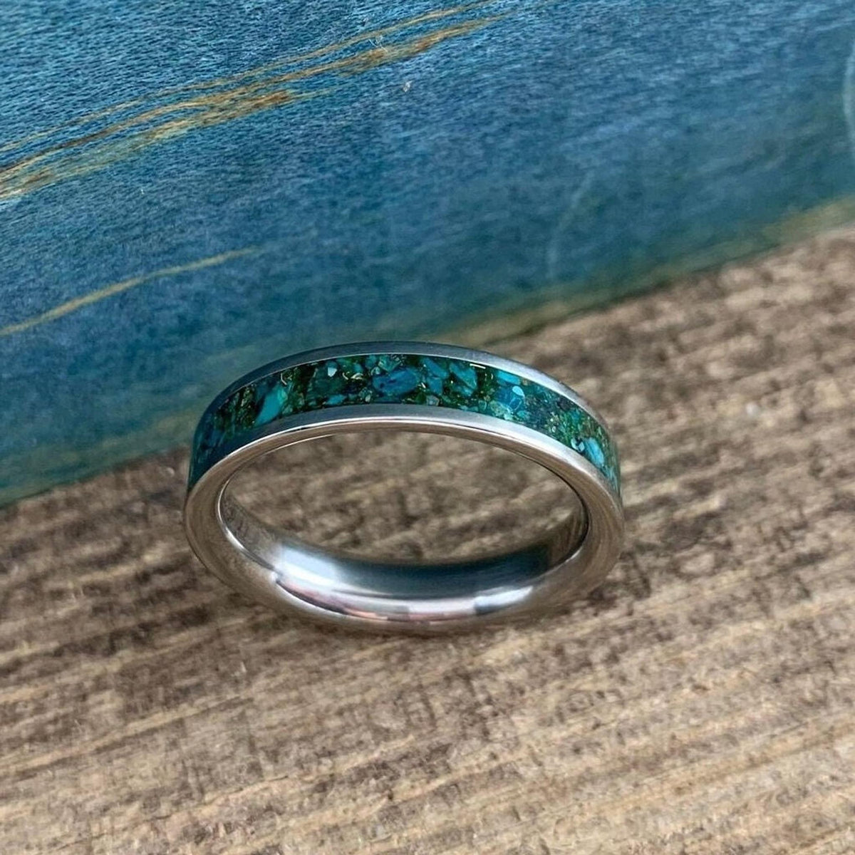 A silver-colored ring with a wide inlay of crushed turquoise and gold-colored flecks, shown head-on on a wooden surface.