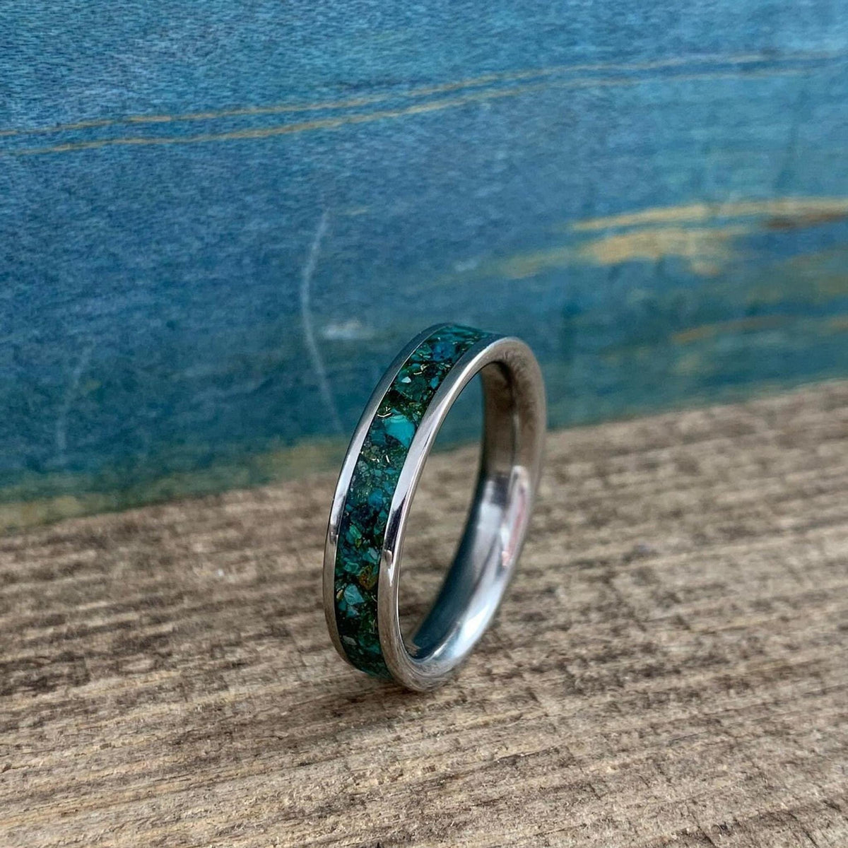 A silver-colored ring with a wide inlay of crushed turquoise and gold-colored flecks, shown at an angle on a wooden surface, with a blue background.