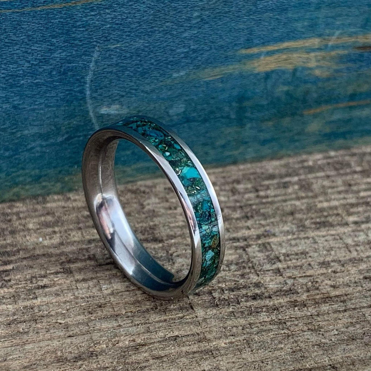 A silver-colored ring with a wide inlay of crushed turquoise and gold-colored flecks, shown at a slight angle on a wooden surface.