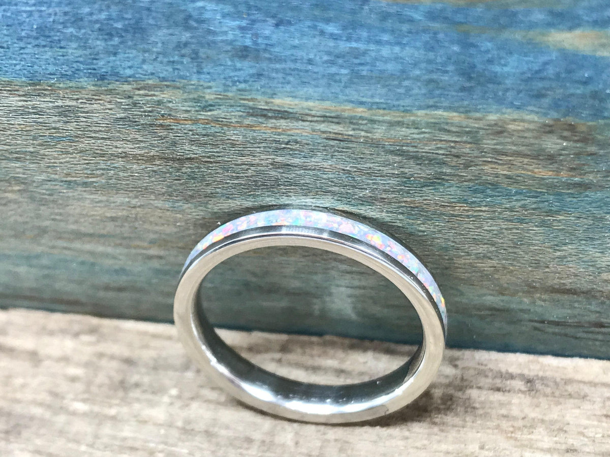 A silver ring with a white opal inlay is shown from a low angle on a light wooden surface, with a blue-painted wood background.