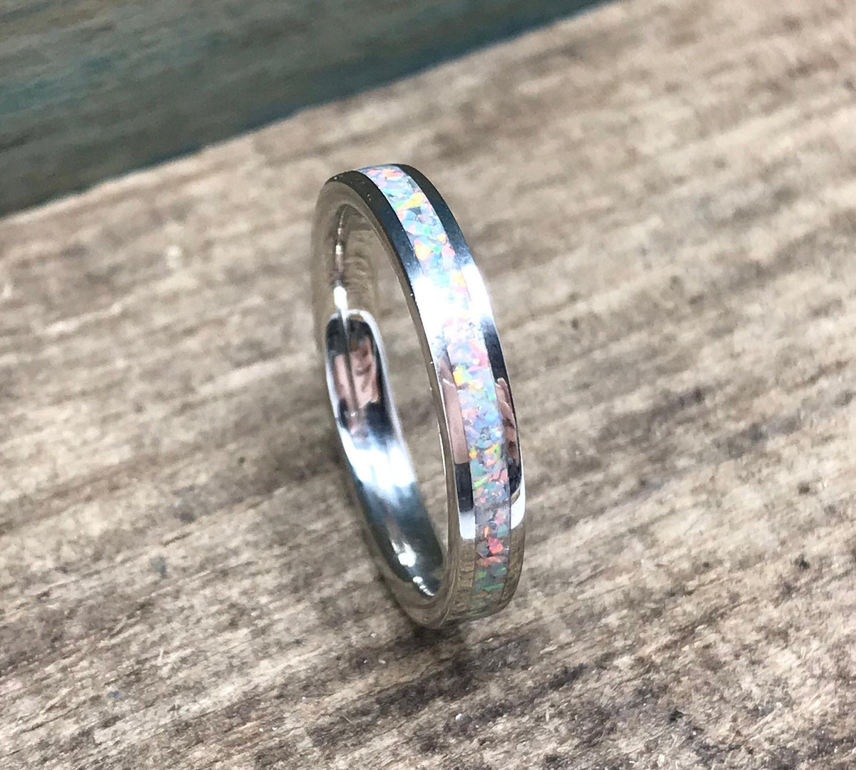 A silver ring with a white opal inlay is centered on a light wooden surface, with a blue-painted wood background.