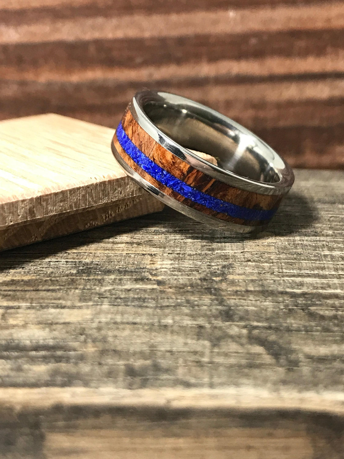A silver-colored ring with a central brown wood band and a thin blue stripe, angled on a wooden surface next to a wood block.