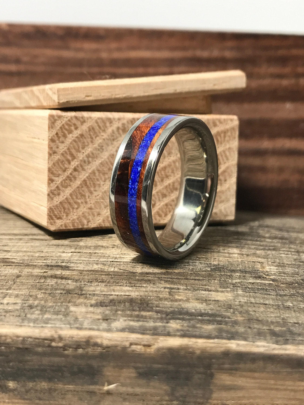 A silver-colored ring with a central brown wood band and a thin blue stripe, resting on a wooden surface with a light wood box behind it.
