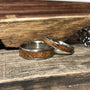 Two silver-toned rings with light brown wood inlays rest on a weathered wooden surface next to a piece of rough-cut wood and a silver chain.