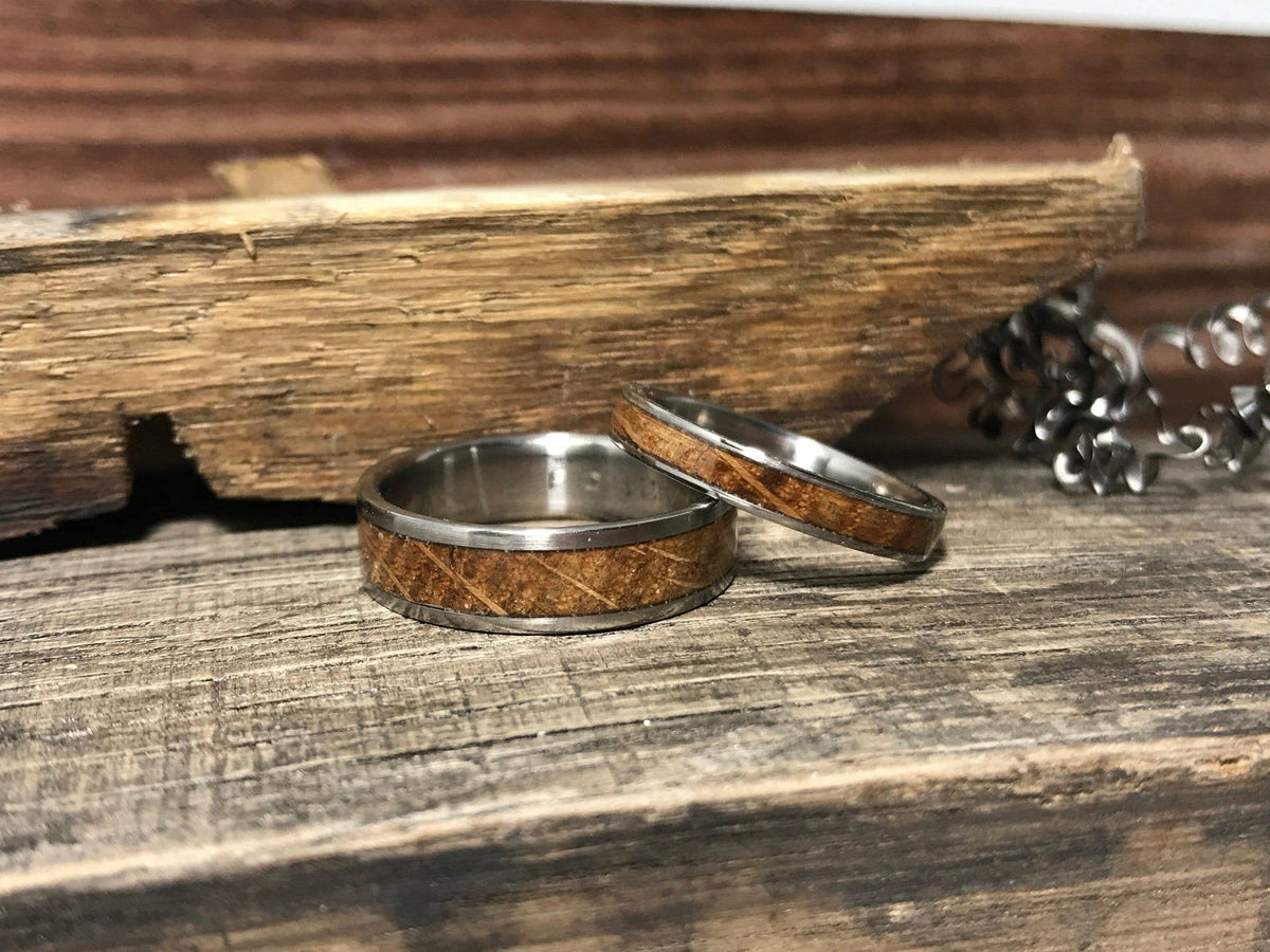 Two silver-toned rings with light brown wood inlays rest on a weathered wooden surface next to a piece of rough-cut wood and a silver chain.
