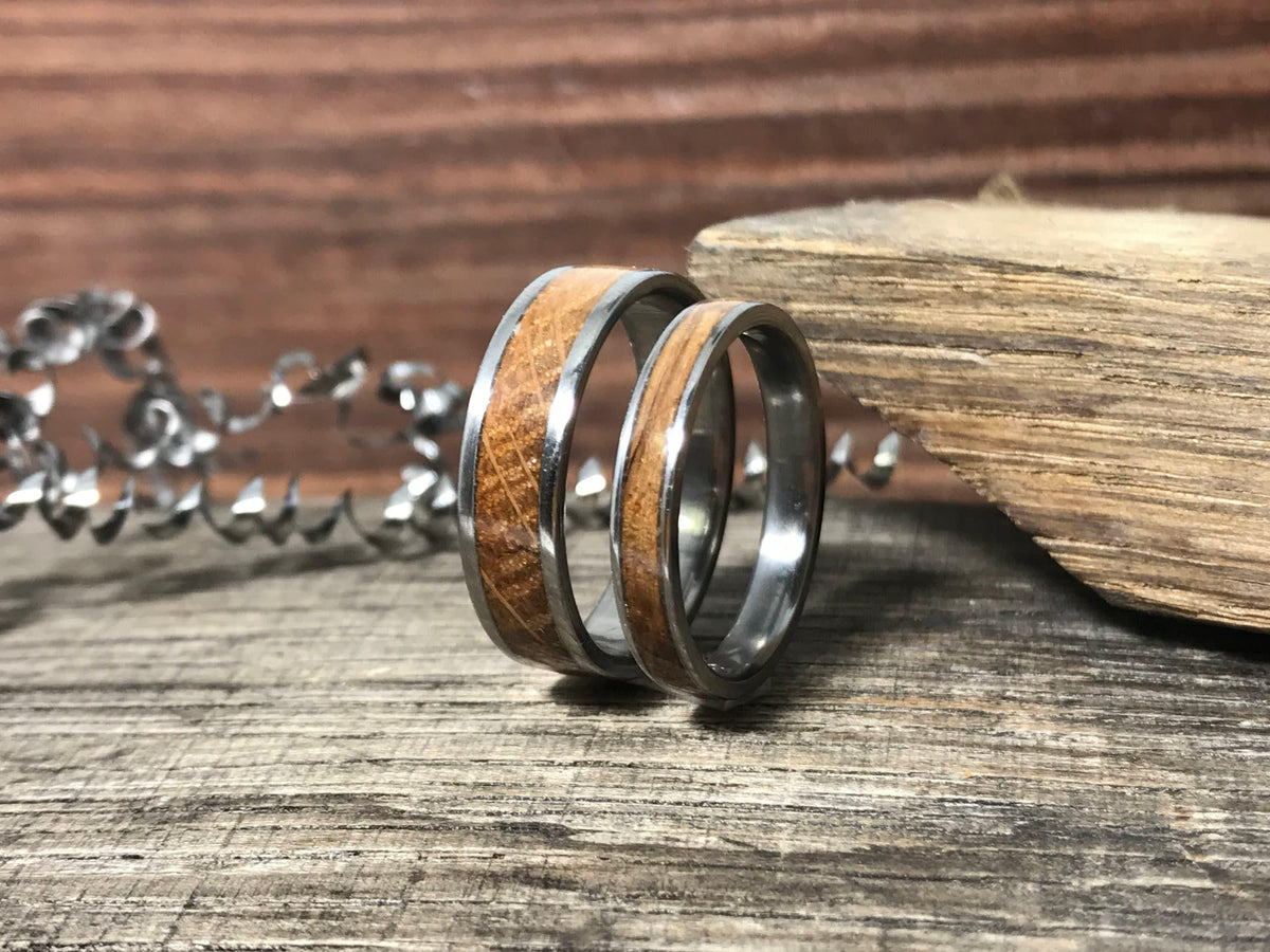 Two silver-toned rings with light brown wood inlays are shown on a weathered wooden surface, with a silver metal shaving spiral and a piece of wood in the background.