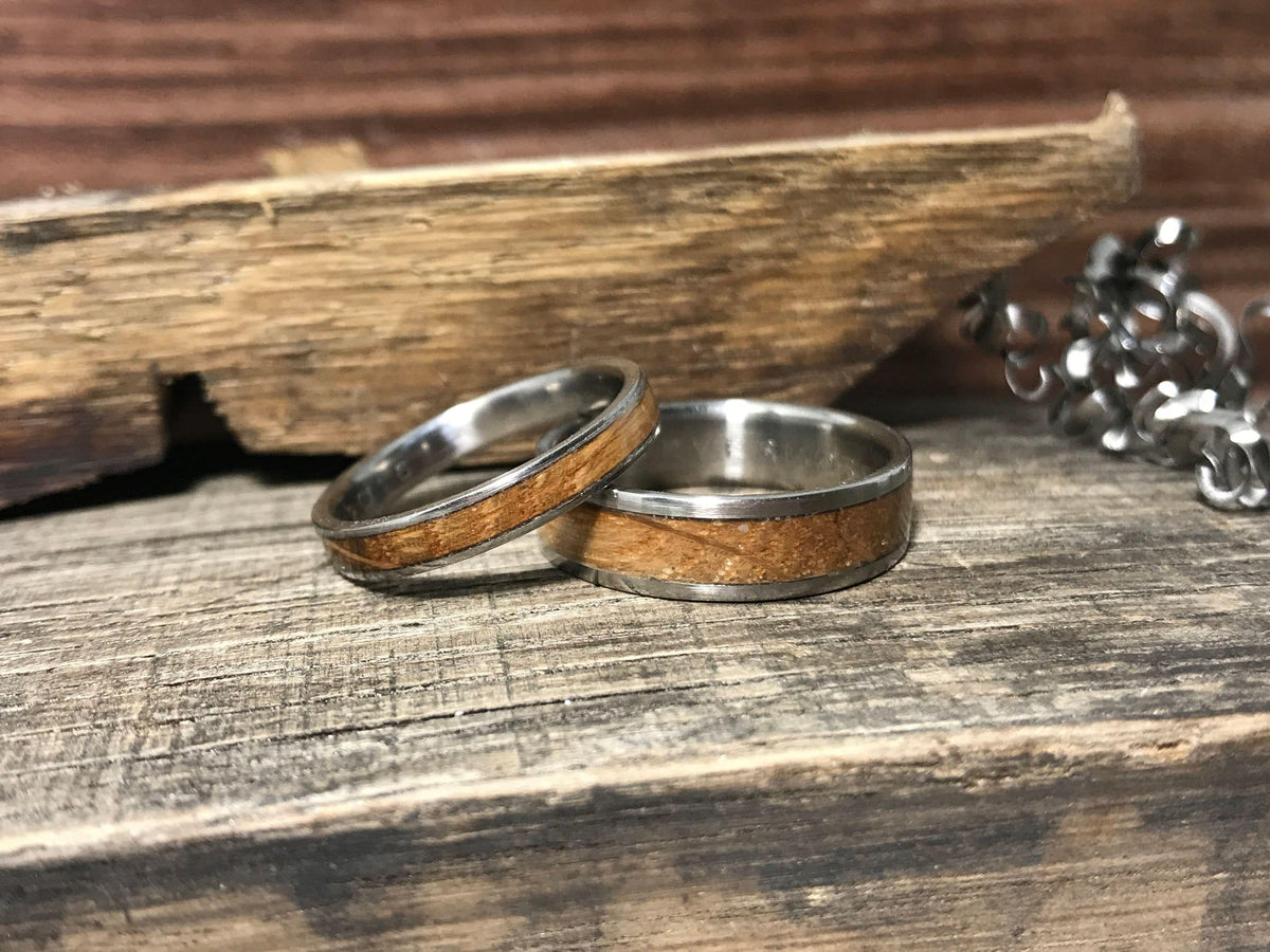 Two silver-toned rings with light brown wood inlays are displayed on a weathered wooden surface, with a piece of rough-cut wood and a silver chain in the background.