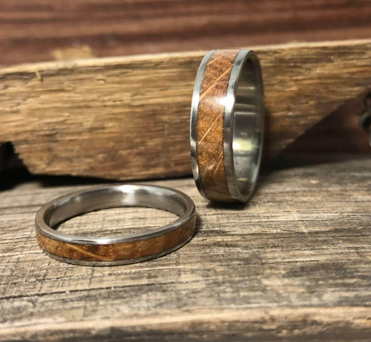 Two silver-toned rings with light brown wood inlays are shown on a weathered wooden surface, with a piece of rough-cut wood in the background.