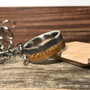A titanium ring with a dark gray and light brown inlay sits on a wooden surface next to metal shavings and a small wooden block.