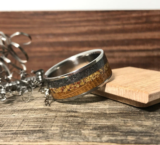 A titanium ring with a dark gray and light brown inlay sits on a wooden surface next to metal shavings and a small wooden block.
