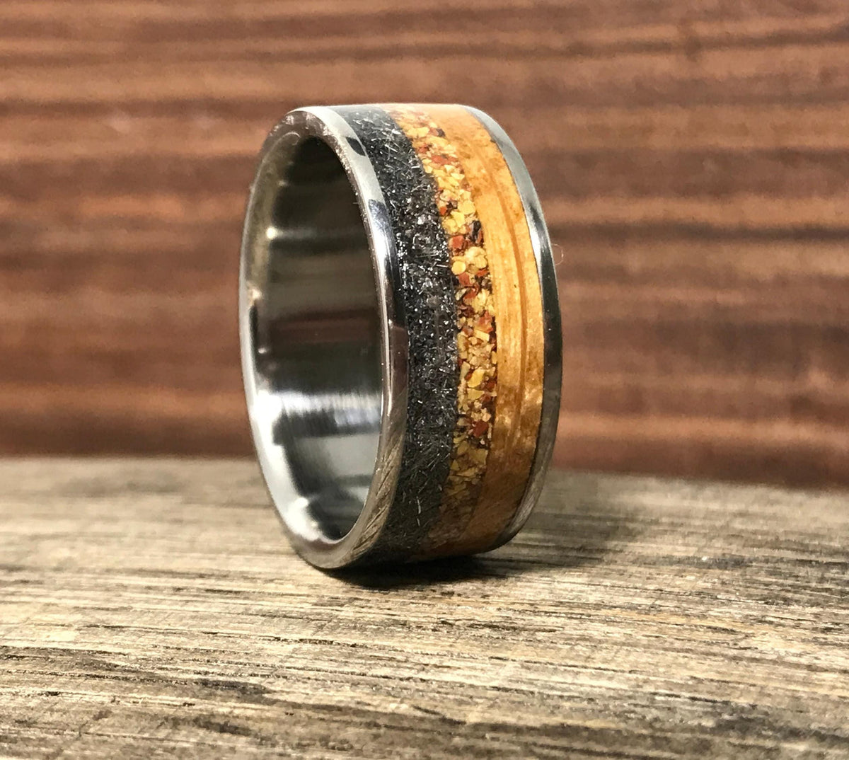 A titanium ring with a dark gray and light brown inlay is shown close-up on a wooden surface, with a blurred wooden background.