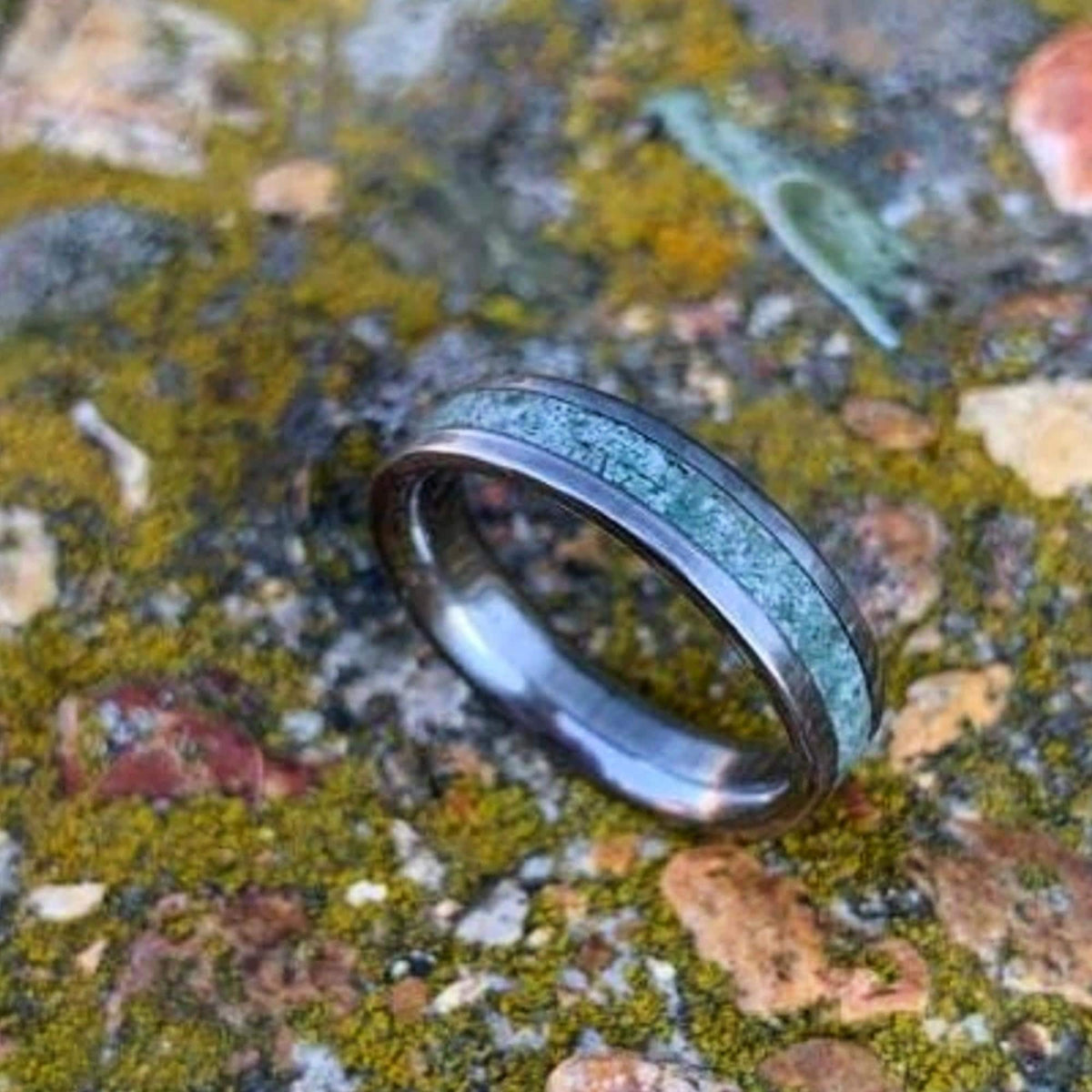 A silver-colored ring with a green moss agate inlay is shown on a mossy rock surface.