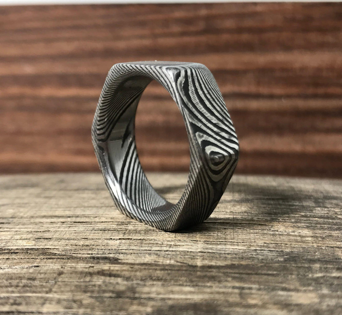 A hexagonal Damascus steel ring with a wavy pattern stands upright on a dark wooden surface, with a blurred wooden background.