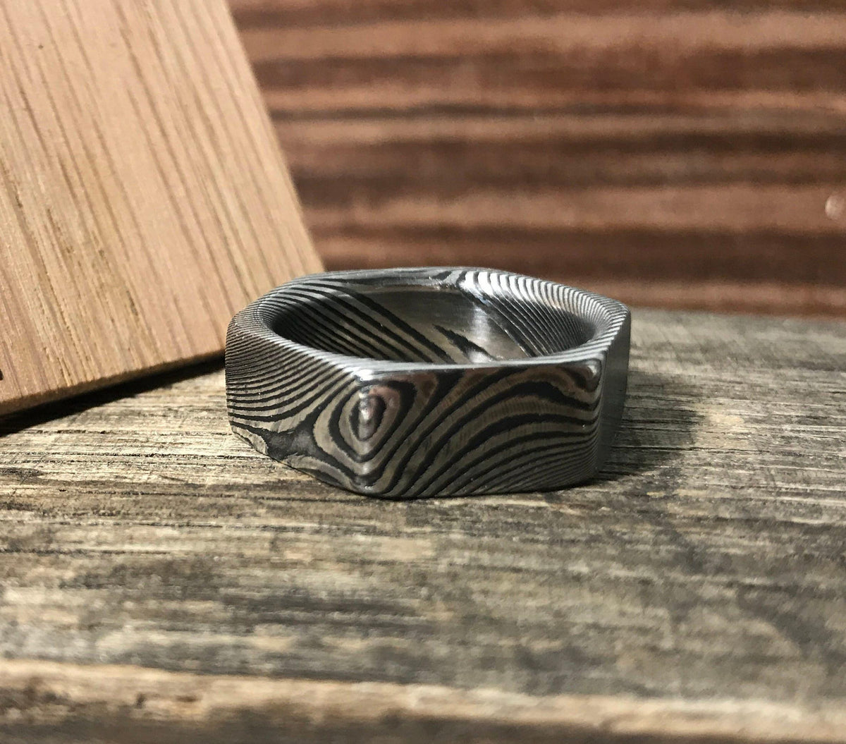 A hexagonal Damascus steel ring with a wavy pattern lies flat on a dark wooden surface, with a light wooden block in the background.