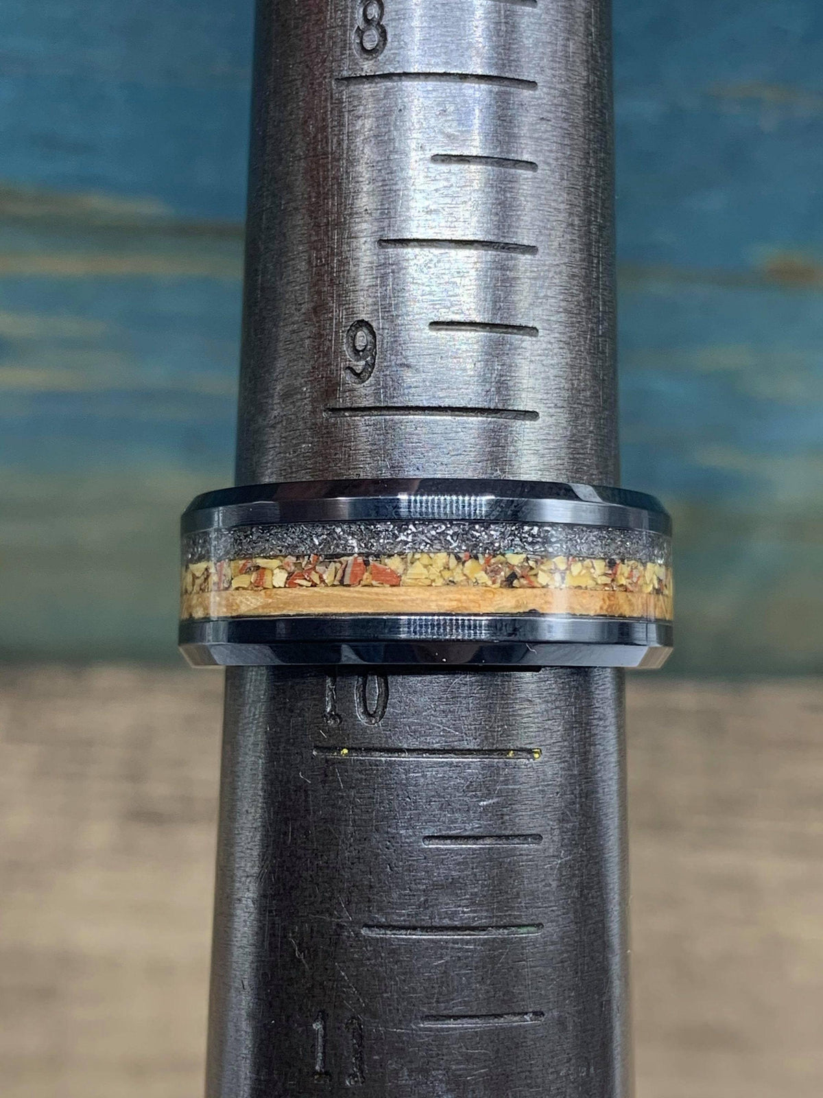 A black ceramic ring with a central inlay of light wood, gray meteorite, and reddish-brown dinosaur bone, displayed on a metal ring sizer.