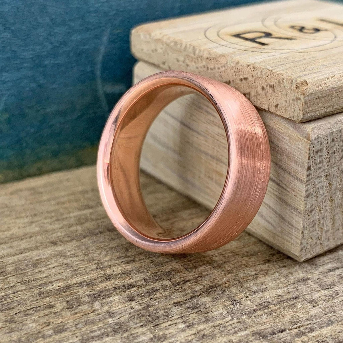 A brushed copper ring with a rounded interior, sitting on a wooden surface next to a light wooden box with "R & B" carved into it.
