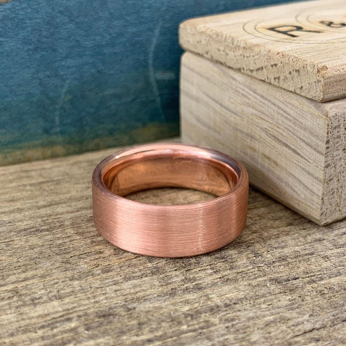 A brushed copper ring with a rounded interior, sitting on a wooden surface next to a light wooden box.