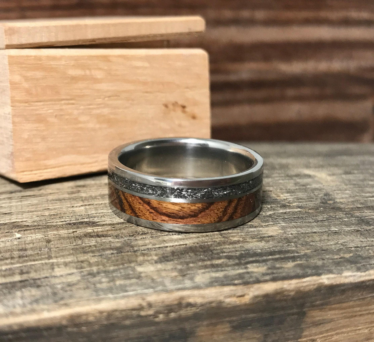 A silver-colored ring with a central wood grain band and a thin, textured gray band, shown from a slightly different angle on a wooden surface.
