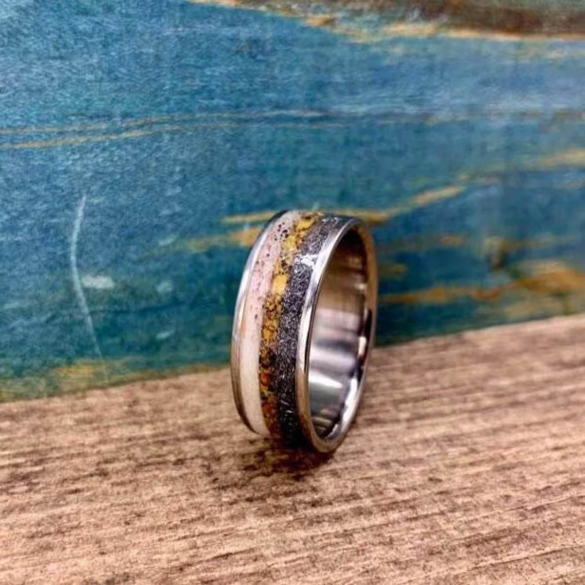 A silver ring with a wide inlay of white, yellow, and black speckled material, shown at an angle on a wooden surface.