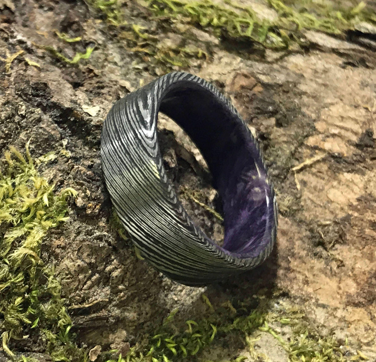 A dark gray Damascus steel ring with a purple interior sits on a mossy log.