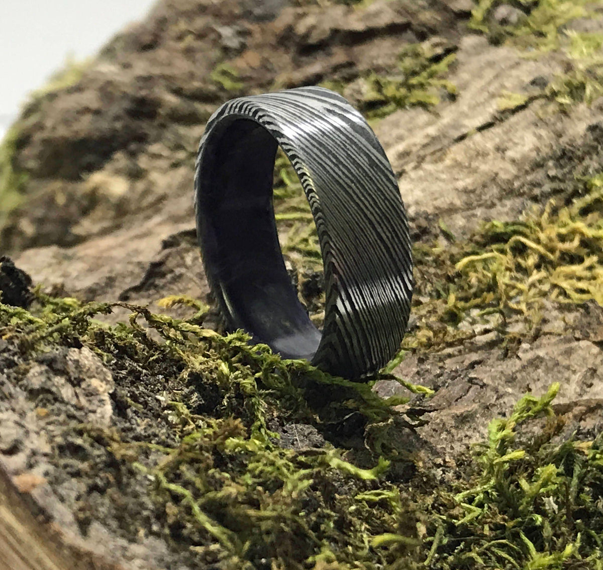 A dark gray Damascus steel ring with a subtle purple interior is placed upright on a mossy log.