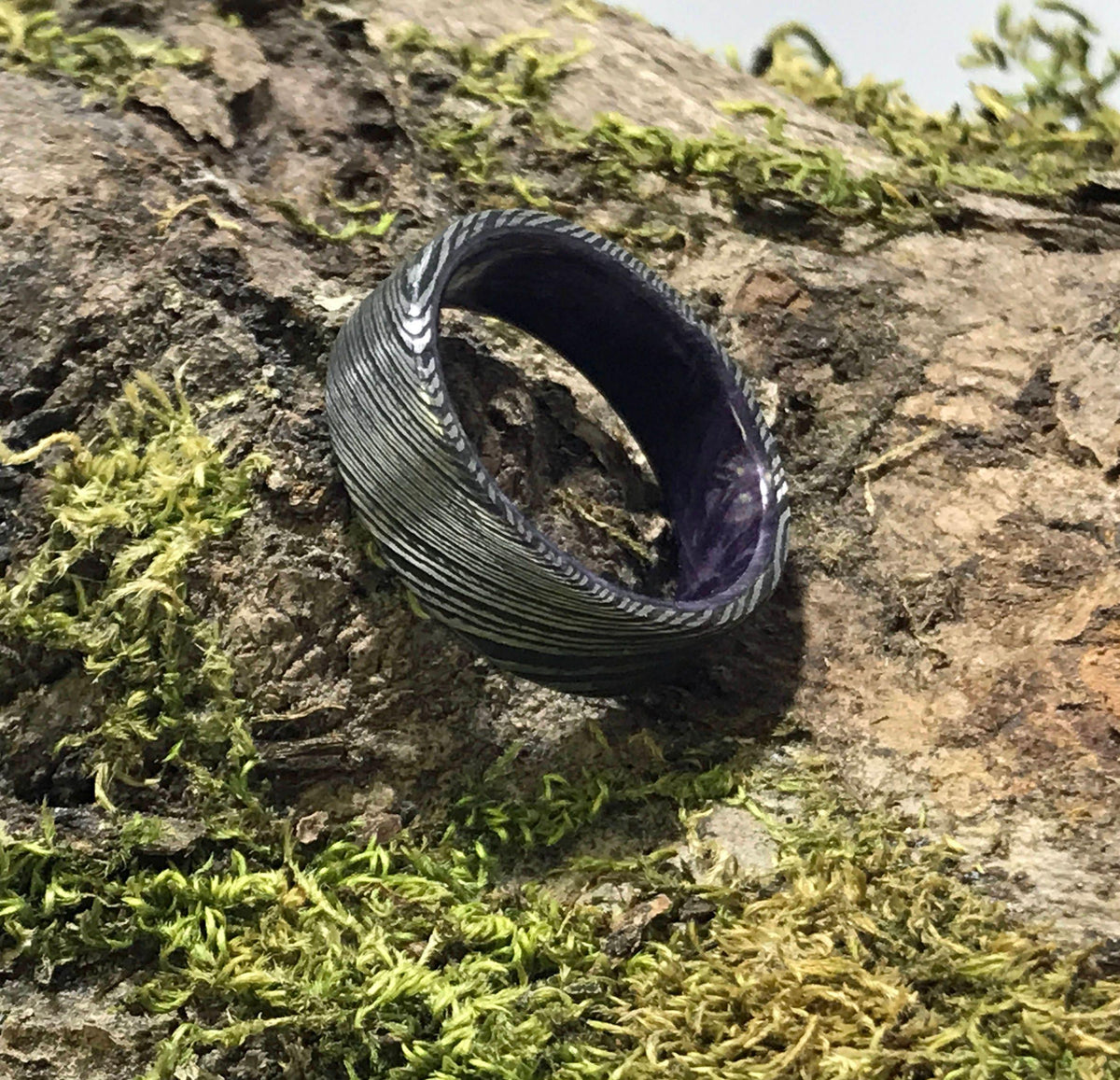 A dark gray Damascus steel ring with a purple interior is shown from a slightly elevated angle on a mossy log.