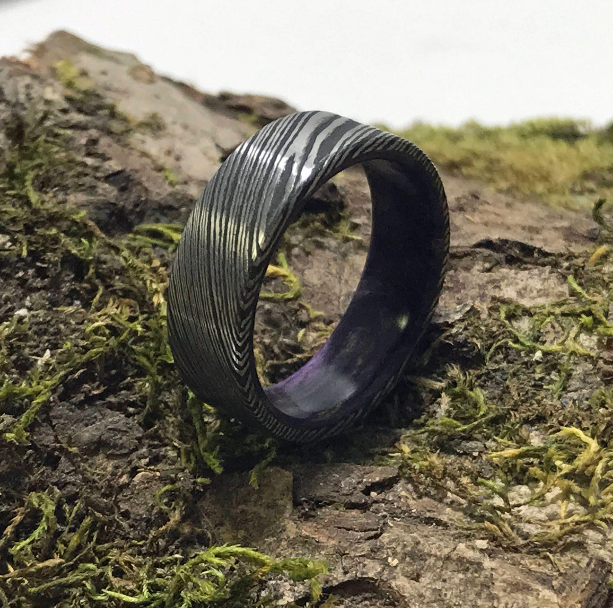 A dark gray Damascus steel ring with a purple interior is positioned on its side on a mossy log.