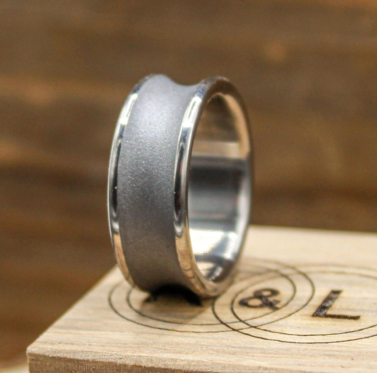 A silver-toned ring with a concave, sandblasted center band and polished edges, resting on a light wood block with a circular logo.