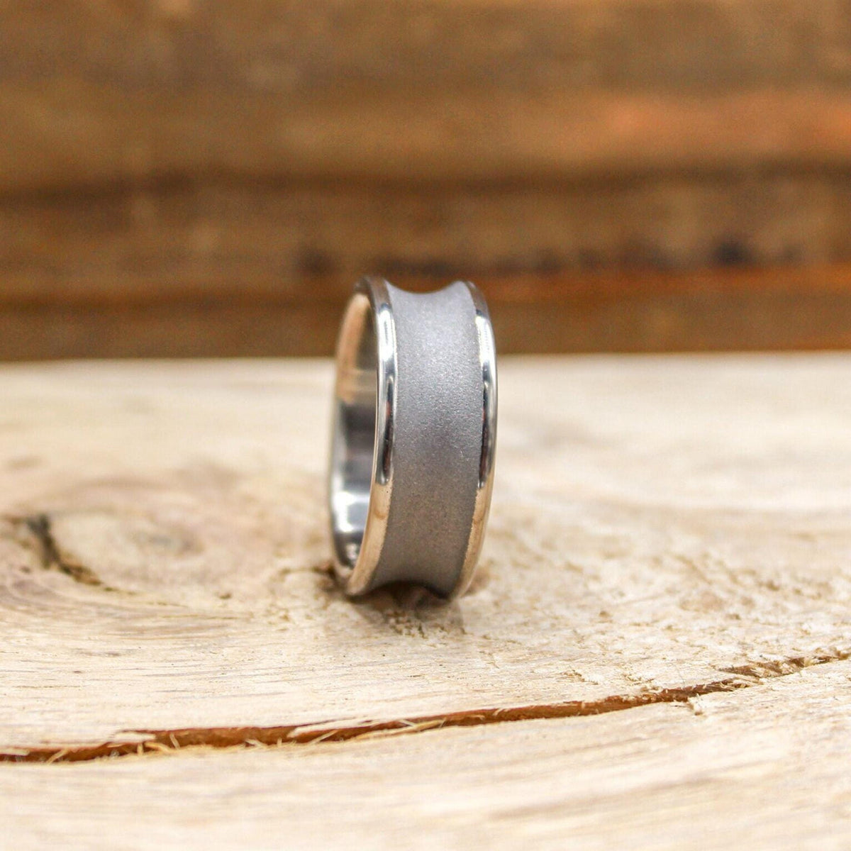 A silver-toned ring with a concave, sandblasted center band and polished edges, viewed from the front on a light wood surface.