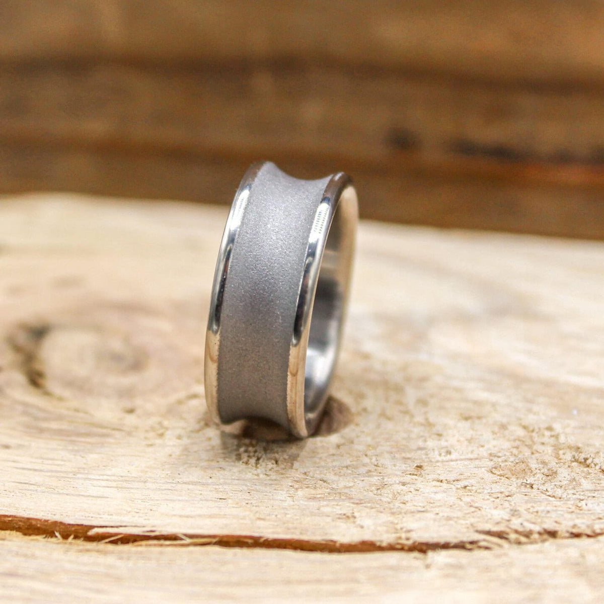 A silver-toned ring with a concave, sandblasted center band and polished edges, viewed at an angle on a light wood surface.