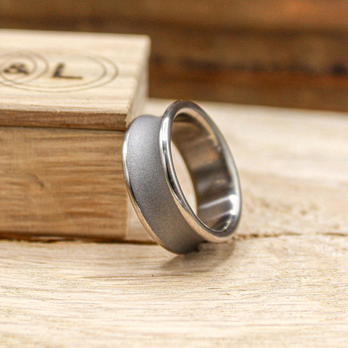 A silver-toned ring with a concave, sandblasted center band and polished edges, leaning against a light wood box.