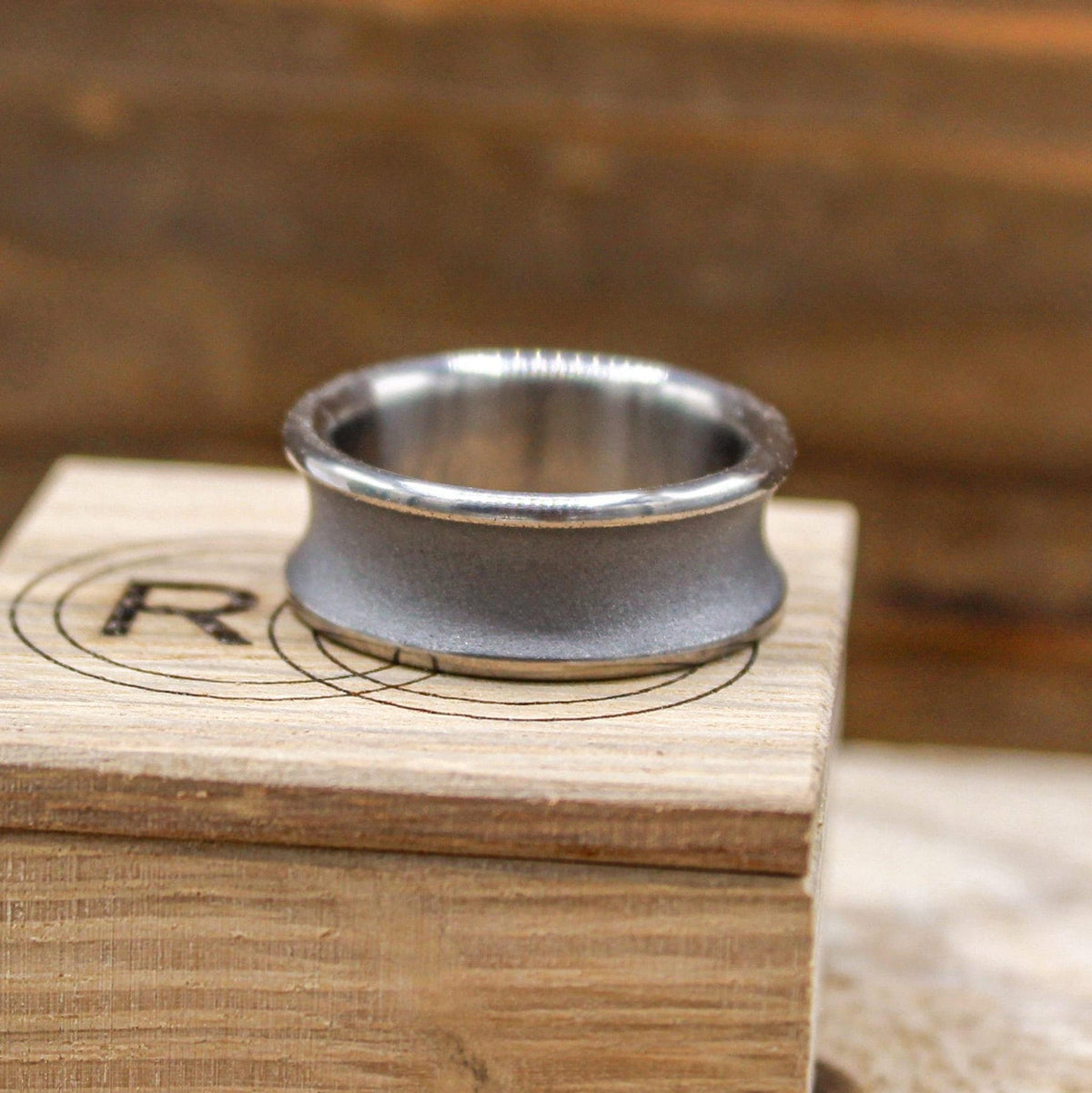 A silver-toned ring with a concave, sandblasted center and polished edges, resting on a light wood block with a circular logo.