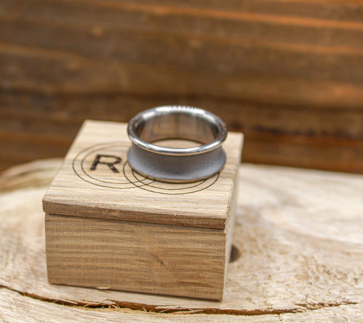 A silver-toned ring with a concave, sandblasted center and polished edges, resting on a light wood box with a circular logo.