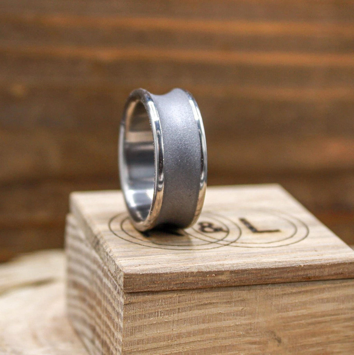 A silver-toned ring with a concave, sandblasted center band and polished edges, viewed at an angle on a light wood block with a logo.