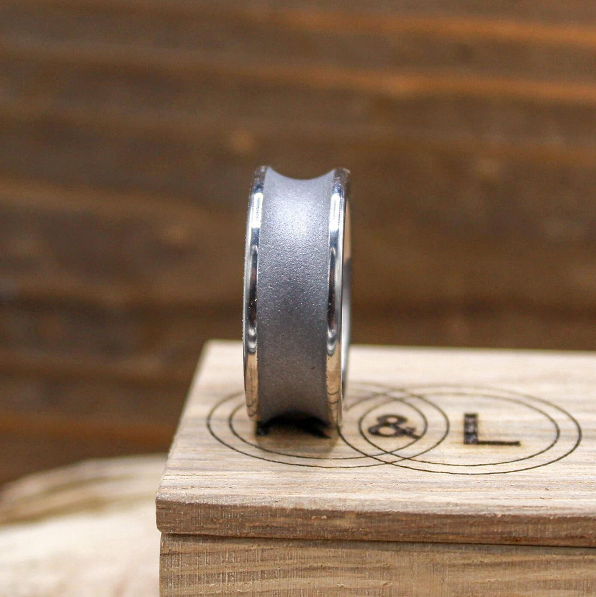 A silver-toned ring with a concave, sandblasted center band and polished edges, viewed from the side on a light wood block with a logo.