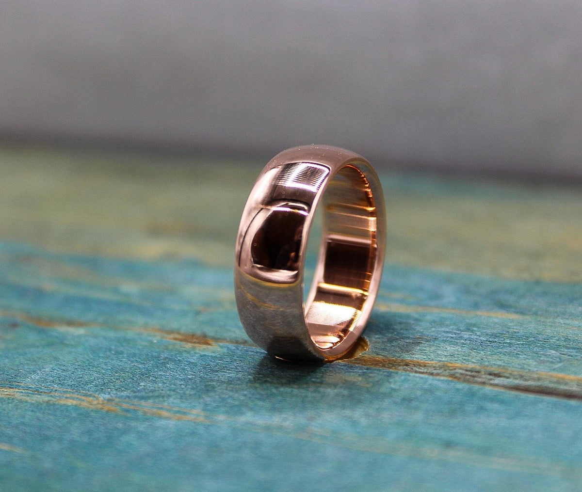 A rose gold-toned ring with a brushed finish on the outer band and a smooth, reflective inner band, resting on a distressed blue-green wooden surface.