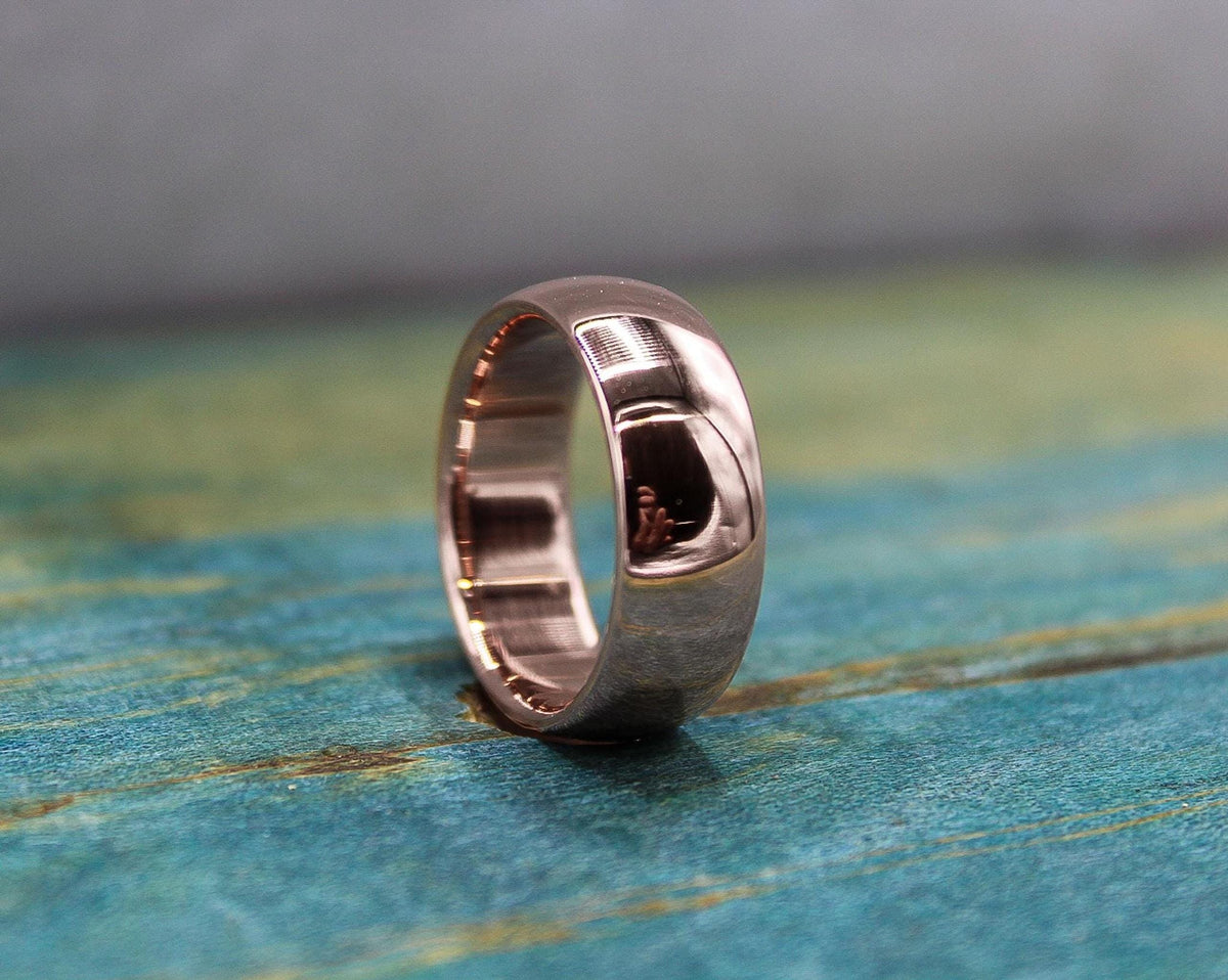 A rose gold-toned ring with a brushed finish on the outer band and a smooth, reflective inner band, resting on a distressed blue-green wooden surface.
