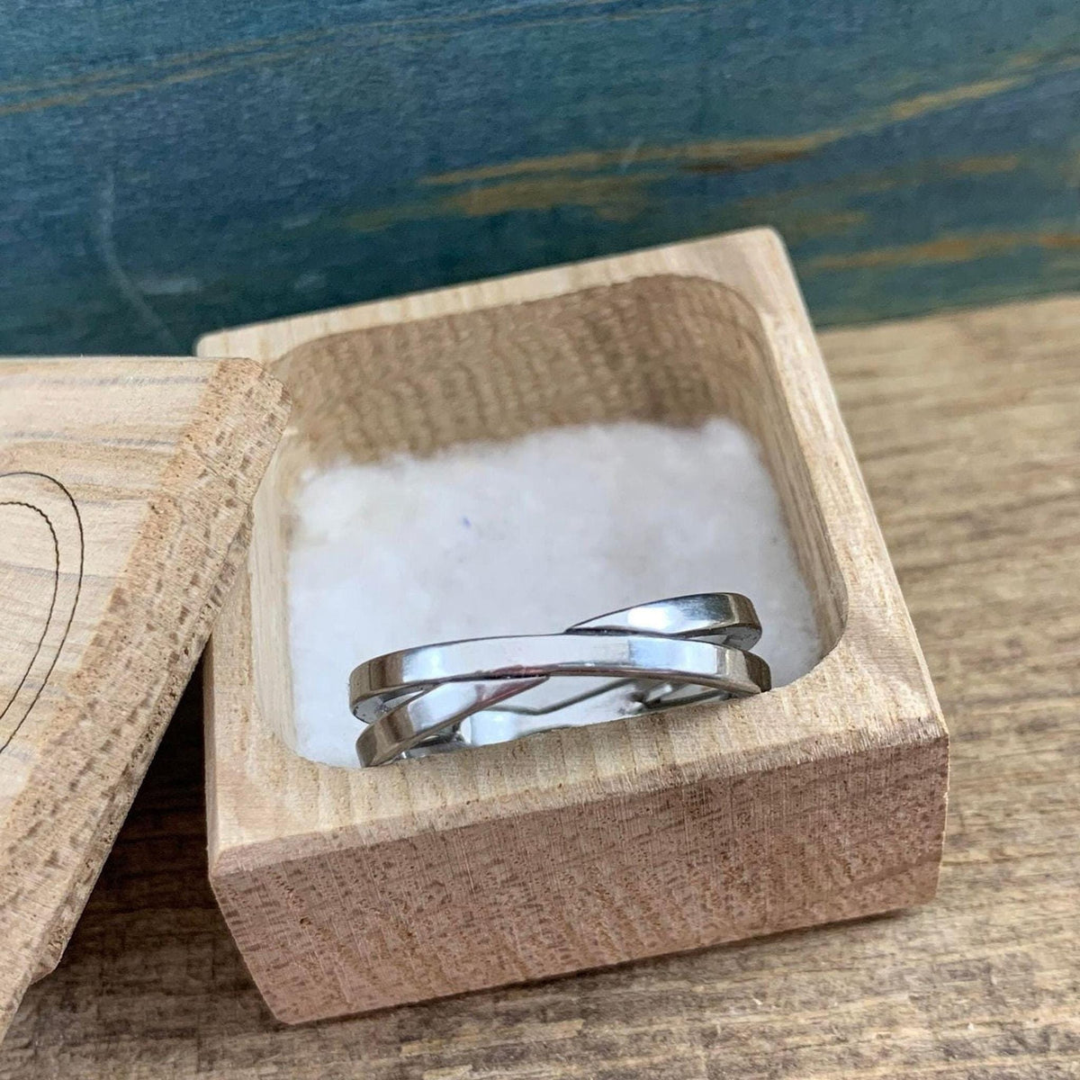 A shiny silver-colored crossover ring with two bands rests on white cotton inside an open light wooden box.