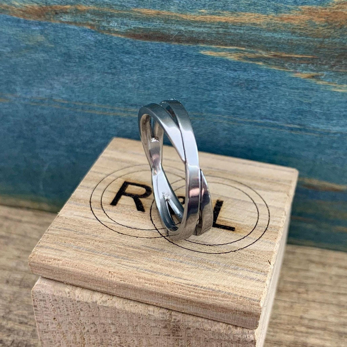 A shiny silver-colored crossover ring with two bands is displayed on a light wooden block with a partially visible "R L" logo, against a blue and brown background.