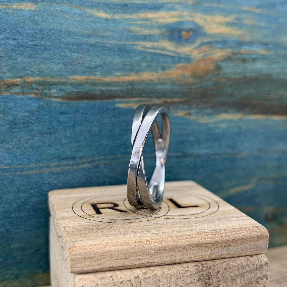 A shiny silver-colored crossover ring with two bands is shown from a slightly angled side view on a light wooden block with a partially visible "R L" logo.